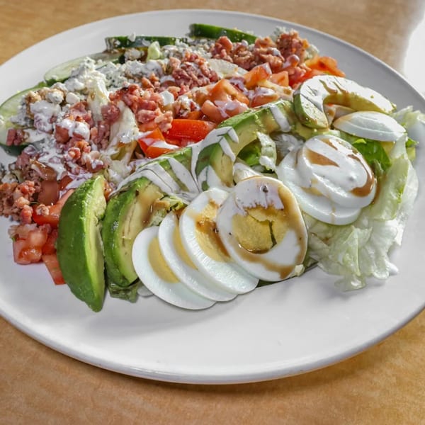 A Cobb salad with avocado, hard-boiled eggs, bacon, tomatoes, and blue cheese on a white plate.