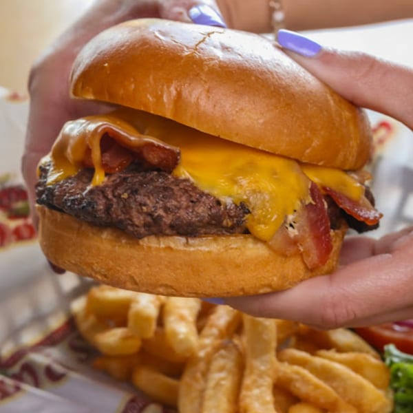 Hands holding a bacon cheeseburger with melted cheese, served with waffle fries at Stevie Tomato's.